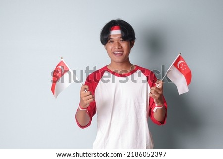 Portrait of attractive Asian man in t-shirt with red and white ribbon on head, holding singapore flag while raising his fist, celebrating success. Isolated image on gray background
