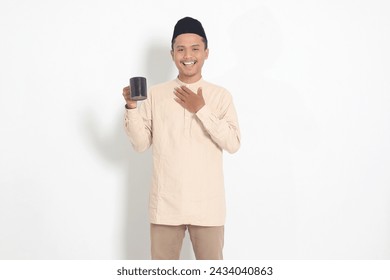 Portrait of attractive Asian muslim man in koko shirt with peci holding a mug, drinking water, quenching thirst after fasting. Isolated image on white background