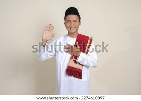 Portrait of attractive Asian muslim man in white shirt with skullcap swears with hands on chest and palms open, makes an oath of allegiance. Isolated image on blue background