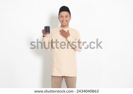 Portrait of attractive Asian muslim man in koko shirt with peci holding a mug, drinking water, quenching thirst after fasting. Isolated image on white background