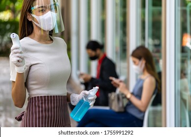 Portrait attractive asian waitress wear face mask and shield hold thermometer and alcohol gel sanitizer with background of social distancing customer waiting in queue. New normal restaurant concept. - Powered by Shutterstock