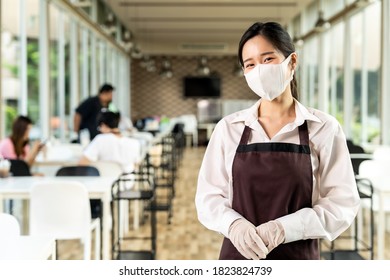 Portrait Attractive Asian Waitress Wear Face With Background Of Customers Sit In Social Distance Table In Restaurant Indoor. New Normal Restaurant Lifestyle Concept.