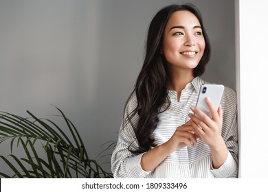 Portrait Of Attractive Asian Woman Looking Outside Window And Smiling Dreamy, Holding Smartphone.