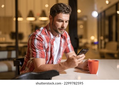 Portrait of an attractive bearded man wearing casual clothes sitting on a couch at the living room, using mobile phone. Handsome millennial man text messaging on smart phone at home.