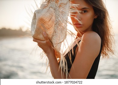 Portrait of attractive blonde girl woman with long hair posing on rocky beach on sunset background,  hold straw hat in her hands and look at the camera