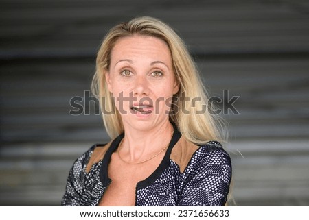 Portrait of an attractive blonde woman with an astonished look teasingly sticks out her tongue, in front of a dark wall