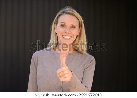 Portrait of an attractive blonde woman giving a thumbs up gesture, in front of wooden wall