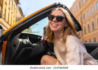 Portrait Of Attractive Blonde Young Woman In Sunglasses, Who Is Looking Out Of A Convertible Car, Smiling Broadly, Shooting From The Bottom Up. Active And Luxurious City Life