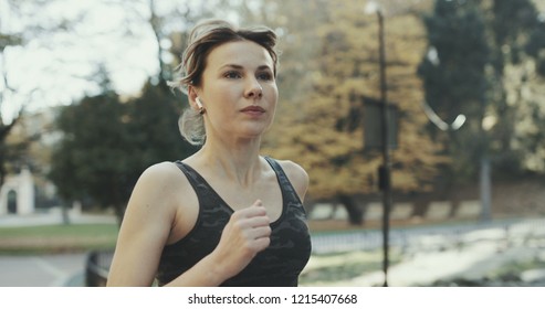 Portrait Of Attractive Brunette Female Runner Running In City Park Airpods Bluetooth Headphones Earphones. Healthy Fitness Athletic Woman Jogging Outdoors