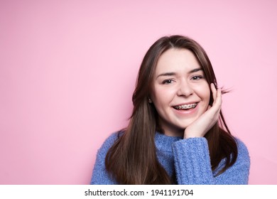 Portrait of an attractive brunette girl in braces, in a blue sweater smiling on a pink background. Dentistry concept, beautiful smile