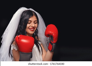 Portrait Attractive Brunette With Red Lipstick, In Wedding Dress, Veil And Boxing Gloves. Young Girl In Role Bride On Black Background, Clasping Her Hands On Her Chest, Protects Herself And Looks Away