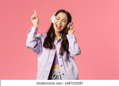 Portrait Of Attractive Carefree Asian Girl Chilling And Vibing, Listening Music In Headphones, Raising Finger Up While Trying Sing Along And Reach Highest Note In Song, Pink Background