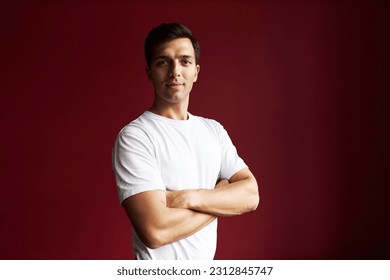 Portrait of attractive caucasian athletic man in white sporty mockup t-shirt posing with folded arms against red studio background, showing his muscular hands and biceps with proud - Powered by Shutterstock