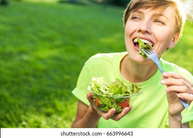 Portrait of attractive caucasian smiling woman eating salad on a sunny summer day sitting on green grass in park, focus on fork with salad - Shutterstock ID 663996643