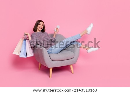 Portrait of attractive cheerful girl in armchair using device holding bags order delivery isolated over pink pastel color background