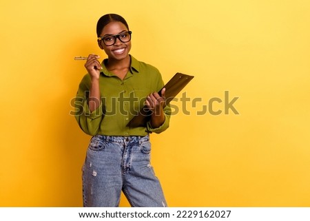 Portrait of attractive cheerful girl holding clipboard signing paper copy space isolated over bright yellow color background