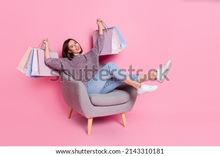 Portrait of attractive cheerful girl sitting in armchair holding bags having fun isolated over pink pastel color background