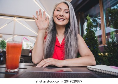 Portrait of attractive cheerful grey-haired woman using laptop talking on zoom waving hi hello at cafe outdoors