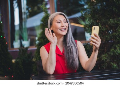 Portrait of attractive cheerful grey-haired woman sitting at cafe using gadget waving hi calling on web cam outdoors