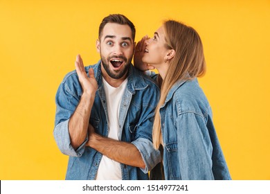 Portrait of an attractive cheerful young couple wearing casual clothing standing isolated over yellow background, telling secrets to each other