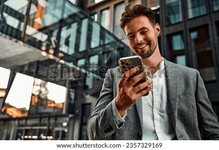 Portrait of attractive confident modern smiling happy adult dressed gray suit man holding a phone and standing outside near office building.  Copy space.                               