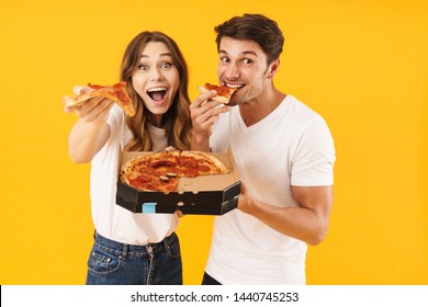 Portrait of attractive couple man and woman in basic t-shirts smiling while eating pizza from box isolated over yellow background