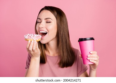 Portrait of attractive dreamy cheerful girl eating doughnut drinking latte isolated over pink pastel color background