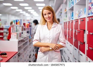 portrait of attractive druggist woman near shelves with medications , posing at camera. smile, wearing white medical uniform