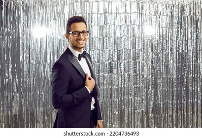Portrait of attractive and elegant showman or host of festive event on silver shiny background. Smiling young dark-skinned man in black classic suit looks confidently at camera. Banner. - Shutterstock ID 2204736493