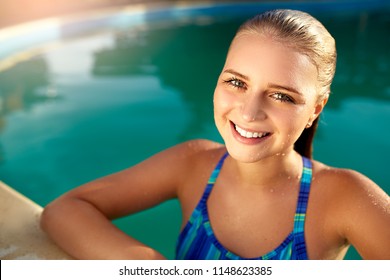 Portrait of attractive emotional blonde girl smiling in the pool side. Young teen wet woman with burning eyes came out of water and looking at camera. Happy tanned female in drops of water swimming.