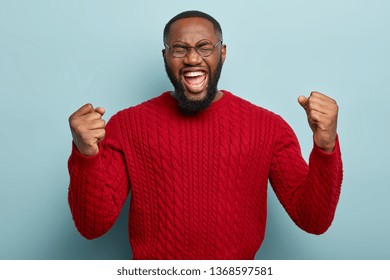 Portrait Of Attractive Emotional Man Shouts For Favourite Team, Clenches Fists, Opens Mouth Widely, Has Eyes Closed, Wears Red Knitted Jumper, Screams, Isolated On Blue. Yeah, Finally Achieved It