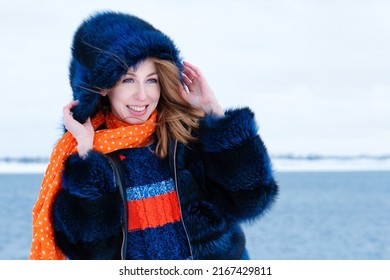 Portrait Attractive Girl Walk In Winter Park On Winter Snow Background. Young Happy Woman In Stylish Warm Outfit And Orange Beanie Hat. Windy Weather