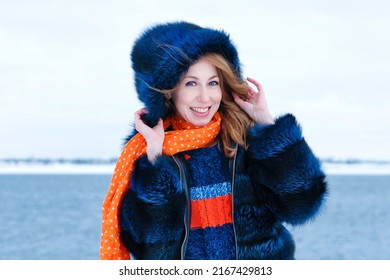 Portrait Attractive Girl Walk In Winter Park On Winter Snow Background. Young Happy Woman In Stylish Warm Outfit And Orange Beanie Hat. Windy Weather