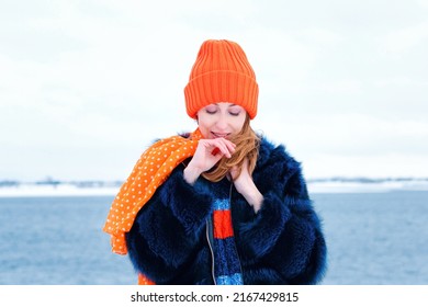 Portrait Attractive Girl Walk In Winter Park On Winter Snow Background. Young Happy Woman In Stylish Warm Outfit And Orange Beanie Hat. Windy Weather