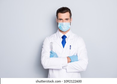 Portrait of attractive handsome dentist with hairstyle in protective face mask, white lab coat, blue tie, holding equipments in crossed arms, isolated on grey background