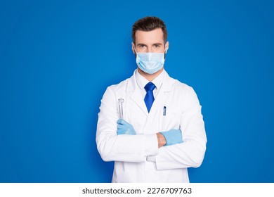 Portrait of attractive handsome dentist with hairstyle in protective face mask, white lab coat, blue tie, holding equipments in crossed arms, isolated on grey background - Powered by Shutterstock