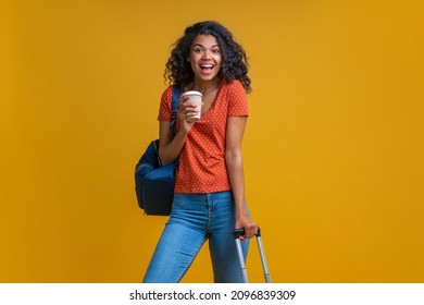 Portrait Of Attractive Happy Smiling Young Tourist Woman Wearing Travel Backpack And  Posing With A Hand Luggage Bag And Paper Cup Of Coffee In Hands, Isolated Over Yellow Background.