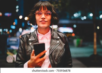 Portrait of attractive hipster girl in eyewear with night light reflection listening to music in earphones standing near copy space area for advertising, young woman enjoying playlist songs on mobile