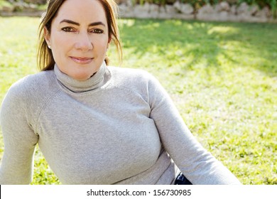 Portrait Of An Attractive Hispanic Middle Aged Mature Woman Relaxing On The Green Grass Of A Home Garden During A Sunny Fall Day, Joyful And Smiling Outdoors.