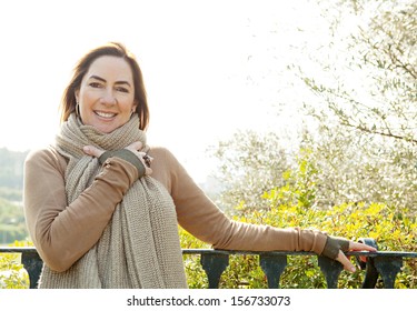 Portrait Of An Attractive Hispanic Middle Aged Mature Woman In A Home Garden Relaxing And Leaning On An Iron Balcony Banister, Looking At Camera And Smiling, Close Up.