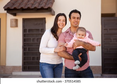 Portrait of an attractive Hispanic young couple and their baby girl standing in front of their new home and smiling