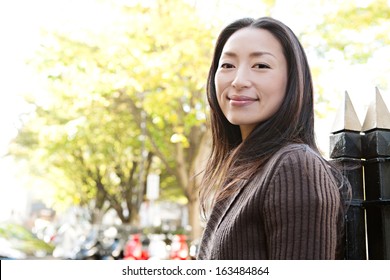 Portrait Of An Attractive Japanese Tourist Woman Visiting The City Of London During A Sunny Day And Standing Near A Park With Black Railings, Smiling And Looking At Camera While Turning, Outdoors.