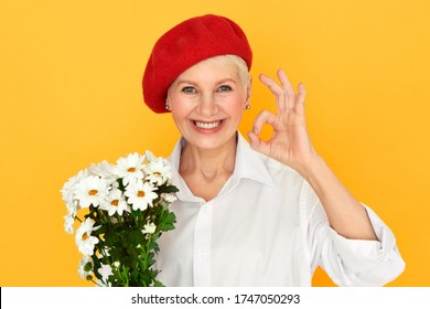 Portrait of attractive joyful middle aged woman florist in red bonnet having confident facial expression, making okay gesture, holding bunch of daisies, arranging flowers for special event