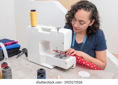 portrait of attractive latin woman using sewing machine, in addition to scissors, threads, fabric, making clothes at home as little businesswoman - Powered by Shutterstock