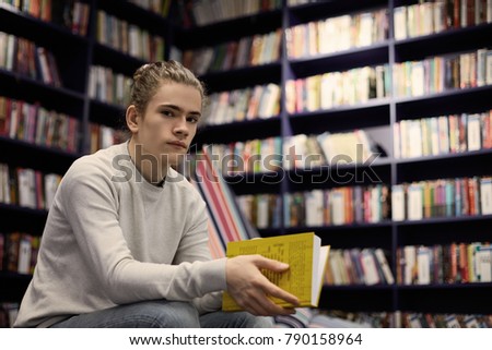 Portrait of attractive male student with hair knot choosing textbook in university library, searching for information for his diploma project, sitting isolated against bookshelves background
