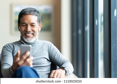 Portrait of attractive mature asian man retired with stylish short beard using smartphone sitting or listening music in urban lifestyle coffee shop. Old man using social network internet technology. - Powered by Shutterstock