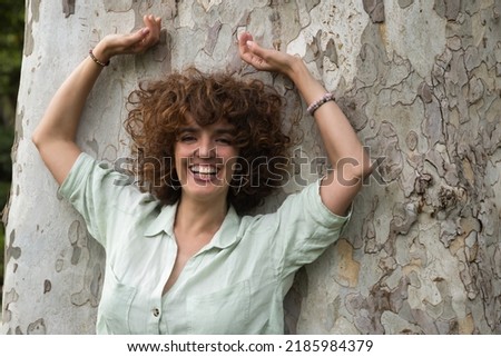 Portrait of attractive mature woman with curly brown hair and shirt, leaning against a tree trunk with her hands on her head laughing happily. Concept happiness, hairstyle, curls, laughter.