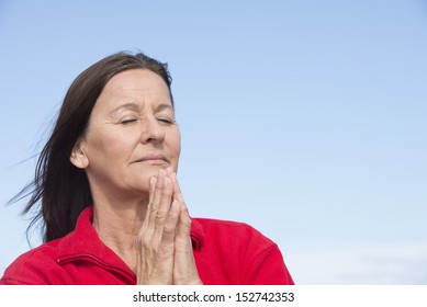 Portrait attractive mature woman relaxed, concentrated, closed eyes, praying, with folded hands at chin, outdoor with blue sky as background and copy space.