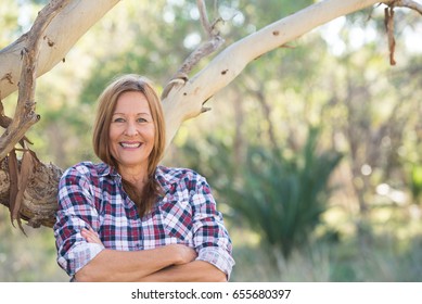 Portrait Attractive Mature Woman In Rural Country, Wearing Plaid Shirt, Posing Relaxed With Happy Smile Against Australian Outback Bush Background.