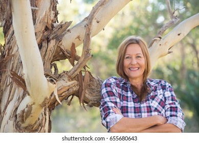 Portrait Attractive Mature Woman In Rural Country, Wearing Plaid Shirt, Posing Relaxed With Happy Smile Against Australian Outback Bush Background.